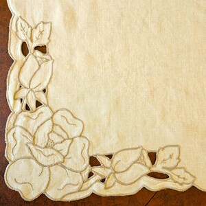 May include: A cream-colored linen tablecloth with a floral cut-out design. The cut-out features roses and leaves with a delicate, embroidered outline. The tablecloth has a scalloped edge and is suitable for a dining table.