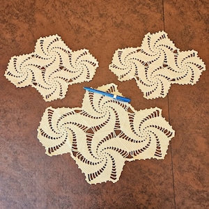 May include: Three cream-colored, star-shaped crochet doilies with a spiral design. A blue pen rests on one of the doilies. The doilies are arranged on a brown surface.