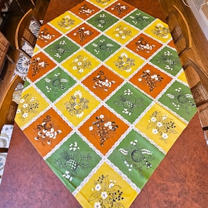 May include: A colorful tablecloth with a diamond pattern featuring floral and fruit designs in yellow, green, and orange. The tablecloth has a white lace trim and is set on a wooden table. The overall theme is vintage kitchen decor.