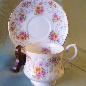 May include: A vintage teacup and saucer set. The white porcelain cup and saucer feature a floral design in pink, yellow, and purple. The set has gold trim and is displayed on a wooden stand. A classic tea set.