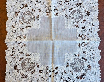 May include: Square white lace handkerchief with intricate floral and scrollwork designs. The delicate lace border frames a plain center, creating a vintage and elegant aesthetic. The handkerchief is displayed on a brown surface.