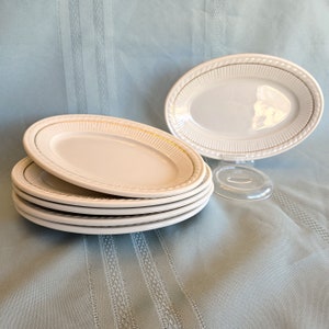May include: A stack of six white oval serving platters with a gold trim and a textured border. One platter is displayed on a clear stand. The platters are on a light blue tablecloth.