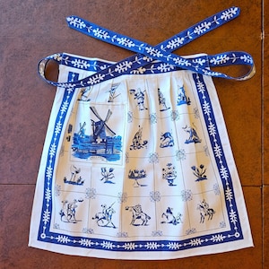 May include: White apron with a blue and white Dutch windmill and folk art design. The apron features a blue border and ties, and is decorated with various blue illustrations. The apron is made of a light-colored fabric.