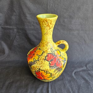 May include: Yellow ceramic vase with a handle and a narrow neck. The vase is decorated with colorful leaf designs in red, orange, and yellow. The vase has a textured surface with brown speckles.