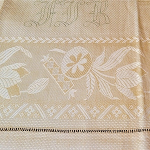 May include: A beige and white woven textile featuring a repeating floral and geometric pattern. The fabric displays an embroidered monogram with the letters FTR at the top, a decorative border with stylized leaves, and a fringed hem edge.