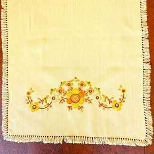 May include: A rectangular, light yellow table runner with a floral embroidery design in shades of yellow, orange, and brown. The edges are finished with a fringe. The embroidered flowers are in the center of the runner.