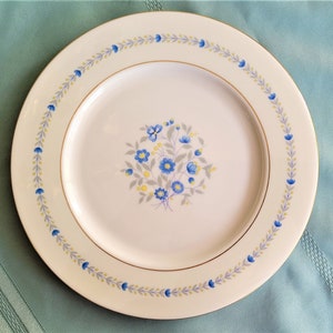 May include: A white ceramic plate with a floral design. The plate features a central bouquet of blue flowers with gray and yellow accents. A border of blue flowers and gray leaves encircles the plate, with a gold rim. The plate is approximately 10 inches in diameter.