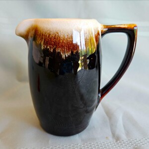 May include: A ceramic pitcher featuring a glossy finish with a dark brown base that transitions into a lighter, creamy white glaze at the top rim. The vessel includes a curved handle on the right side and a small pouring spout on the left side.