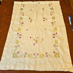 May include: Cream-colored rectangular tablecloth with embroidered floral design. The embroidery features purple flowers, green vines, and yellow heart-shaped leaves. The tablecloth is laid out on a dark brown wooden surface.