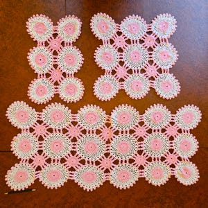May include: Handmade crochet doilies in a soft pink and cream color scheme. The set includes three pieces: two smaller square-shaped doilies and one larger rectangular doily. The doilies feature a floral design with a central pink circle.