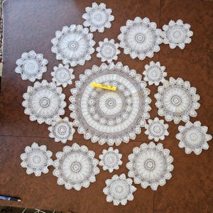 May include: A collection of white, intricately crocheted doilies in various sizes, arranged on a brown surface. The doilies feature detailed floral and circular patterns. A yellow utility knife rests on the largest doily, providing scale.