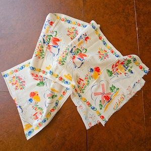 May include: Cream-colored vintage Dutch-themed fabric with a repeating pattern of windmills, houses, and figures in traditional attire. The fabric is bordered with a floral design in red, yellow, and blue.