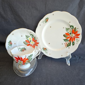 May include: A white porcelain teacup, saucer, and plate set with a Christmas poinsettia design. The set features red, white, and green floral patterns with gold trim and star accents. The plate measures approximately 23 cm in diameter.