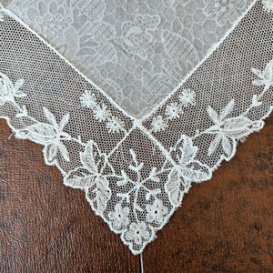 May include: Close-up of a white lace handkerchief. The handkerchief features a delicate floral pattern and a detailed, openwork border. The lace is finely woven, creating a textured appearance. The handkerchief is a classic accessory.