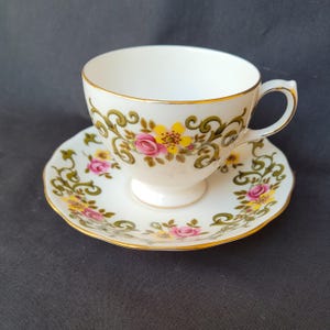 May include: White teacup and saucer set with a floral design. The cup and saucer have a gold trim and feature pink and yellow flowers with green accents. The teacup has a handle and sits on a small pedestal.