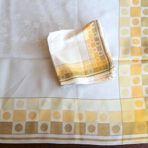 May include: A white tablecloth with a floral pattern and a matching stack of napkins. The border and napkin trim feature a geometric design of yellow squares and circles, with a brown accent.