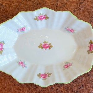 May include: Oval-shaped, white ceramic serving dish with a scalloped edge and a light green trim. The dish is decorated with pink rose floral patterns. A decorative piece for serving food.