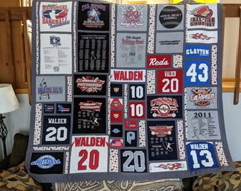 May include: A handmade baseball-themed quilt with various patches featuring team names, numbers, and tournament details. The quilt showcases a mix of colors, including red, white, blue, and gray, with text like "WALDEN" and numbers like "20" and "43".