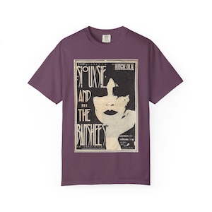 May include: Plum-colored t-shirt featuring a vintage-style concert poster for Siouxsie and the Banshees. The poster is in black and white, with the band's name prominently displayed above an illustration of a woman's face. The shirt has short sleeves and a crew neck.