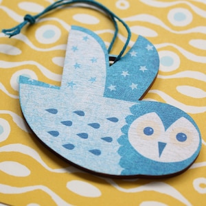 May include: A wooden owl decoration in light blue and white. The owl features a blue body with white star patterns on the wings and teardrop shapes on the chest. A blue string is attached for hanging.