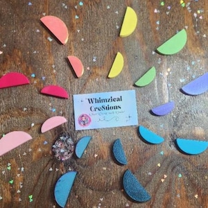 May include: A collection of small, semi-circular pieces in various colors including pink, red, yellow, green, blue, and purple, arranged around a rectangular business card that reads Whimzieal CreStions with a small circular logo and decorative stars.
