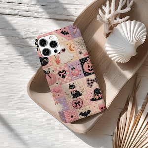 May include: A patchwork phone case featuring Halloween-themed designs. The case has a variety of squares with images of ghosts, black cats, bats, pumpkins, and bows in pink, purple, and black. The phone case is on a wooden tray with a seashell and coral.