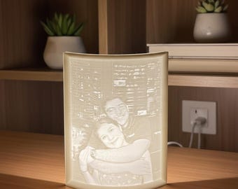 May include: A cream-colored, curved-edge lamp with a photo of a couple embracing, set against a cityscape backdrop. The lamp is illuminated, showcasing the image. The lamp is on a wooden surface, with a bookshelf and plants in the background.