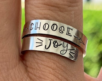 May include: A silver ring with the words "Choose Joy" engraved on it. The ring is worn on a person's finger.