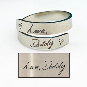 May include: A silver ring with the words "Love, Daddy" engraved on it. The ring is a simple band with a heart symbol on each side of the inscription.
