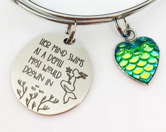 May include: A silver bangle bracelet with a charm that reads "Her mind swims at a depth most would drown in" with a mermaid illustration and a green and blue heart charm with mermaid scales.