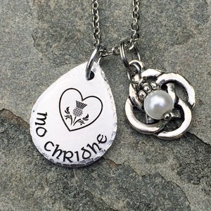 May include: A silver necklace with a teardrop-shaped pendant that says "mo chridhe" and features a heart with a stylized thistle inside. The necklace also has a charm with a Celtic knot design and a pearl in the center.
