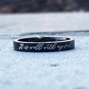May include: A black ring with the inscription "It is well with my soul" engraved in white lettering.