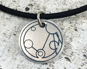 May include: A silver necklace with a black cord and a round pendant featuring a stylized design of circles and lines.