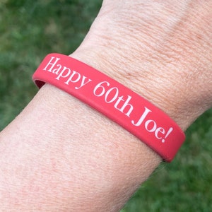 May include: A red silicone wristband with the text "Happy 60th Joe!" in white. The wristband is worn on a wrist, with a green grassy background.