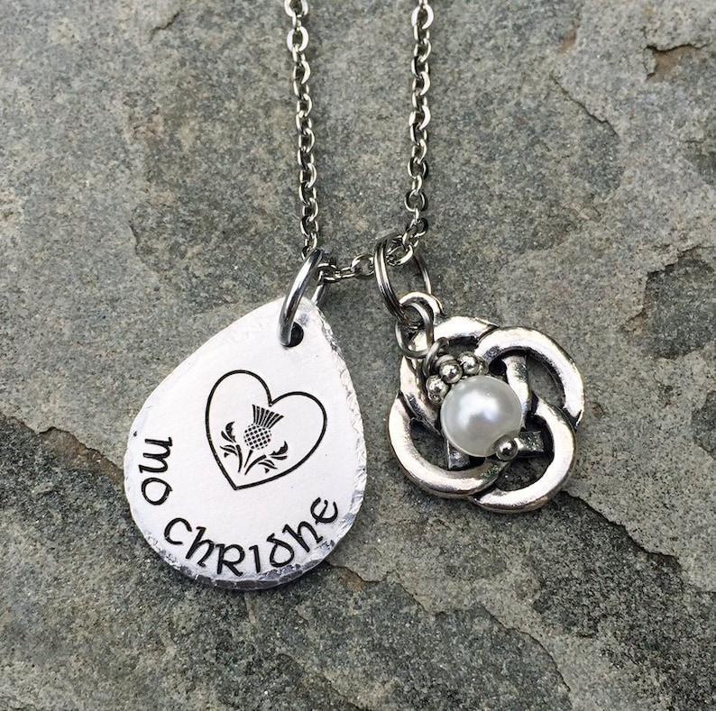 May include: A silver necklace with a teardrop-shaped pendant that says "mo chridhe" and features a heart with a stylized thistle inside. The necklace also has a charm with a Celtic knot design and a pearl in the center.