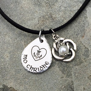 May include: A silver necklace with a heart-shaped pendant featuring a thistle and the text "mo chridhe". The necklace also has a Celtic knot charm with a pearl.