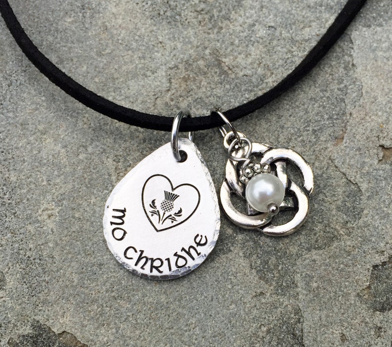 May include: A silver necklace with a heart-shaped pendant featuring a thistle and the text "mo chridhe". The necklace also has a Celtic knot charm with a pearl.