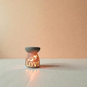 May include: A ceramic candle holder with a gray top and a pink base. The base has heart-shaped cutouts and the word "LOVE" in a decorative font. A warm light shines from within, casting shadows.