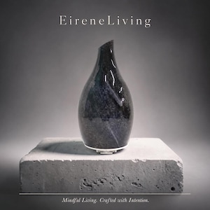 Puede incluir: Un jarrón decorativo azul oscuro y gris con una forma asimétrica única, colocado sobre un bloque de hormigón gris. El texto "EireneLiving" está encima del jarrón, con "Mindful Living. Crafted with Intention." debajo.
