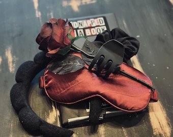 May include: A dark red satin sleep mask with a black fork-shaped accessory and a dark red rose. A black scrunchie and a black padded headband are also visible. The book in the background has the text "Kidnapped by a Book".