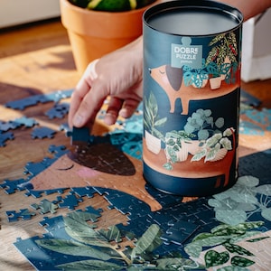 Könnte beinhalten: Eine dunkelblaue zylindrische Puzzlebox mit einer Dackel-Illustration und dem Text "DOBRE PUZZLE JAMNIK" liegt neben einem teilweise fertigen Puzzle. Das Puzzle zeigt ein Hunde- und Pflanzenmotiv. Eine Hand legt ein Puzzleteil.