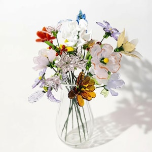 Puede incluir: Un jarrón de cristal transparente lleno de un colorido ramo de flores artificiales. Las flores son de varios colores, incluyendo blanco, naranja, amarillo, morado y marrón. Las flores están hechas de un material translúcido.