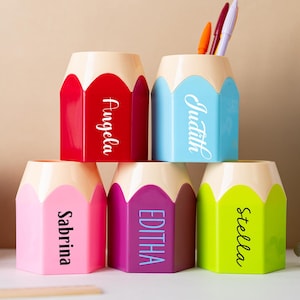 Puede incluir: Un conjunto de portalápices con forma de lápiz de colores brillantes: rojo, azul, rosa, morado y verde. Cada portalápices tiene un nombre escrito en escritura blanca. Algunos contienen bolígrafos y lápices.