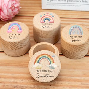 Puede incluir: Cajas de madera para guardar dientes de leche con diseños de arcoíris y el texto "MILK TEETH FROM" seguido de un nombre. Las cajas cilíndricas tienen un acabado de madera natural. Una caja está abierta.