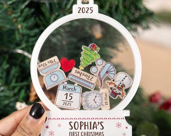 Op de afbeelding: Een witte kerstbal met een helder midden, gevuld met kleine, kleurrijke stickers. De ornament heeft de tekst "2025" bovenaan en "SOPHIA'S FIRST CHRISTMAS" onderaan. Stickers zijn onder meer een weegschaal, hart, liniaal en sneeuwpop.
