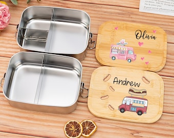 May include: Two stainless steel lunch boxes with dividers. The lunch boxes are on a wooden surface with two personalized wooden lids. The lids feature a food truck design with the names "Olivia" and "Andrew" written on them.