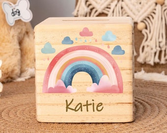 May include: Wooden rainbow piggy bank with a personalized name "Katie" on the front. The rainbow is painted in shades of pink, blue, and yellow with white clouds and raindrops.