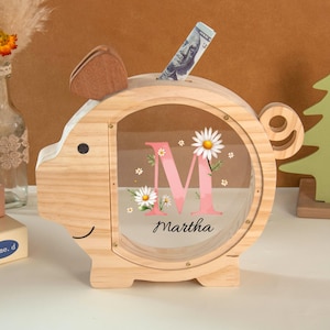 May include: A wooden piggy bank shaped like a pig, with a clear front panel. The panel features a pink floral "M" and the name "Martha". A dollar bill is inserted into the slot. The bank has a wooden ear and tail.