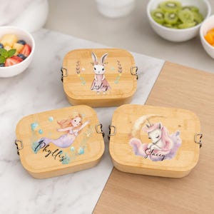 Puede incluir: Tres fiambreras rectangulares de bambú con cierres metálicos. Una muestra un conejo y el nombre Lesley, otra una sirena y el nombre Phyllis, y la tercera un unicornio sobre una luna creciente con el nombre Stacey impreso en las tapas.