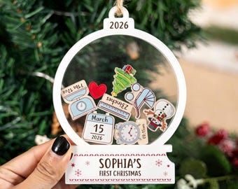 May include: A white, circular, clear-fronted Christmas ornament featuring the year 2026 at the top. Inside are small icons of a scale labeled 71bs 12oz, a heart, a tree, a ruler labeled 23inches, a stroller, a snowman, and a calendar showing March 15 2026.
